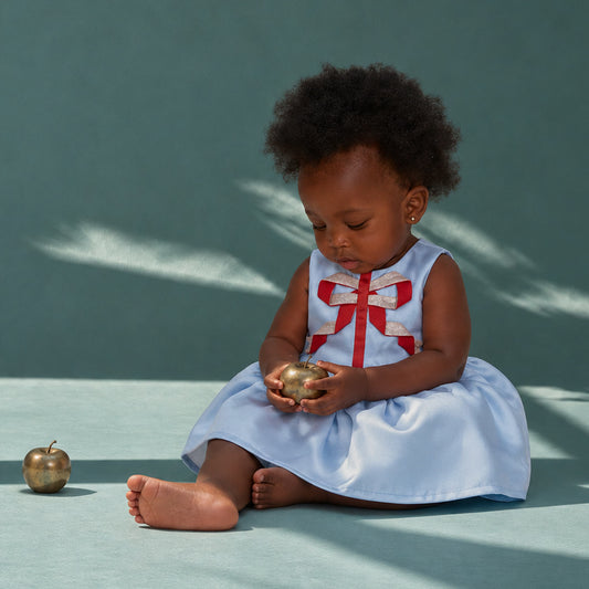 A toddler girl sits barefoot on a teal studio floor, wearing a sleeveless light blue dress with a large red bow at the neckline. She holds a small metallic gold object in her hands, with a matching gold apple prop on the floor beside her.