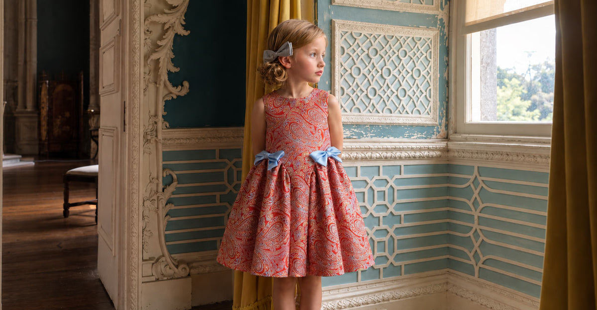A young girl with blonde hair in a grey bow stands in an ornate room wearing a sleeveless coral pink patterned dress with blue bows at the waist.