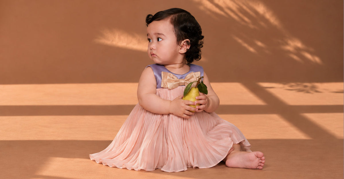 A baby girl wearing a sleeveless pleated dress with a lavender bodice and a large gold bow sits barefoot on a warm brown studio floor, holding a green pear.