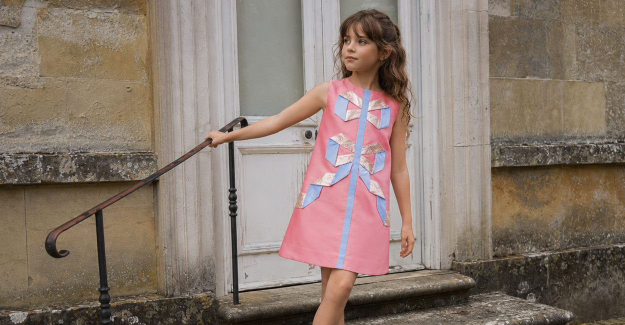 A young girl with long brown hair stands on stone steps in front of a building doorway, wearing a sleeveless pink dress with a large bow applique on the front and a vertical blue stripe down the center. She is holding onto a dark metal handrail.