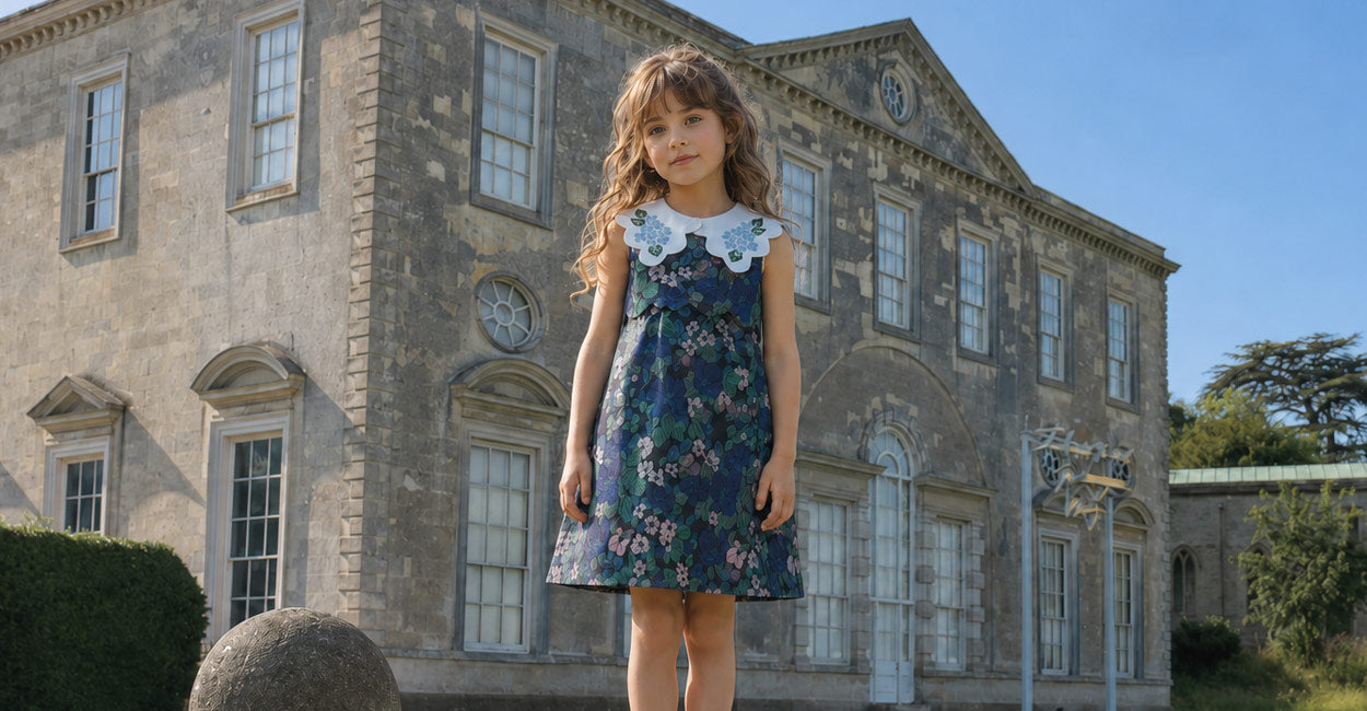 A young girl with long wavy brown hair stands in front of a large historic stone mansion wearing a sleeveless navy floral dress with a white Peter Pan collar.