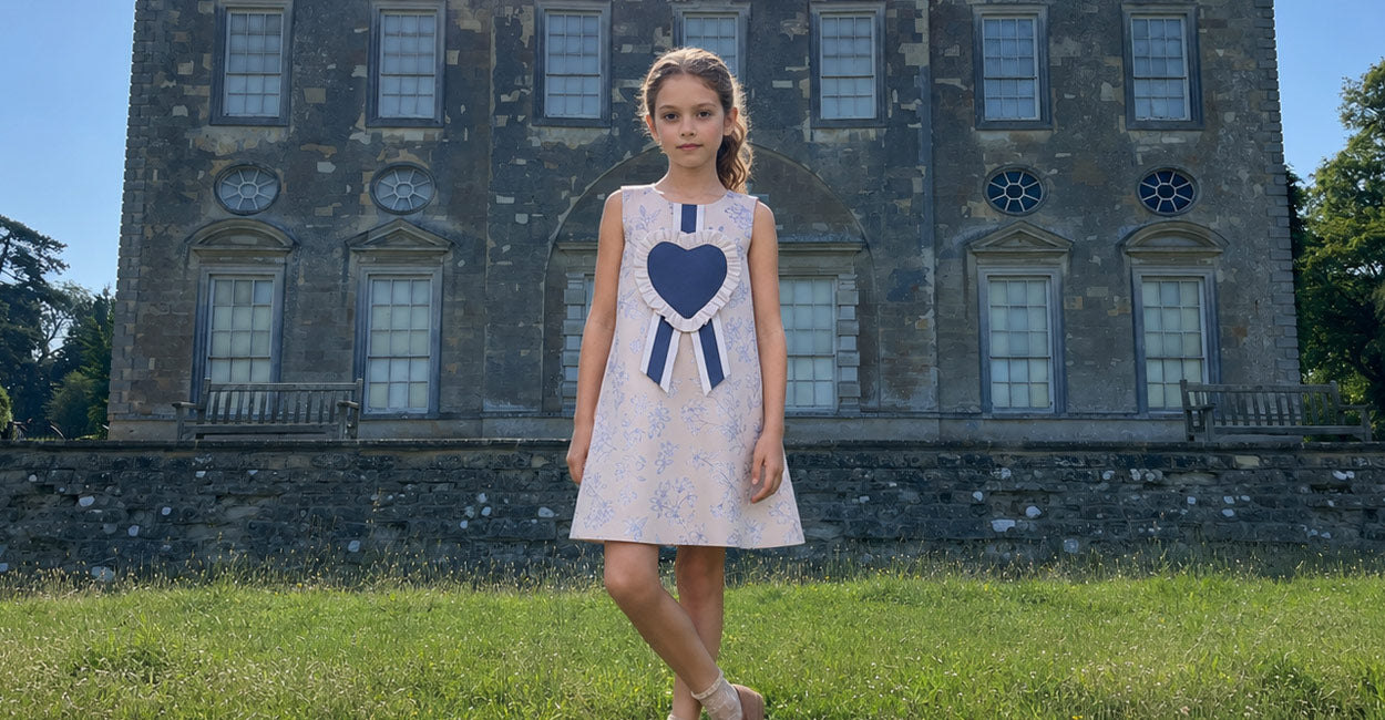 A young girl stands on a grassy lawn in front of a large stone mansion, wearing a sleeveless white dress with a large navy blue heart and ribbon motif on the front.
