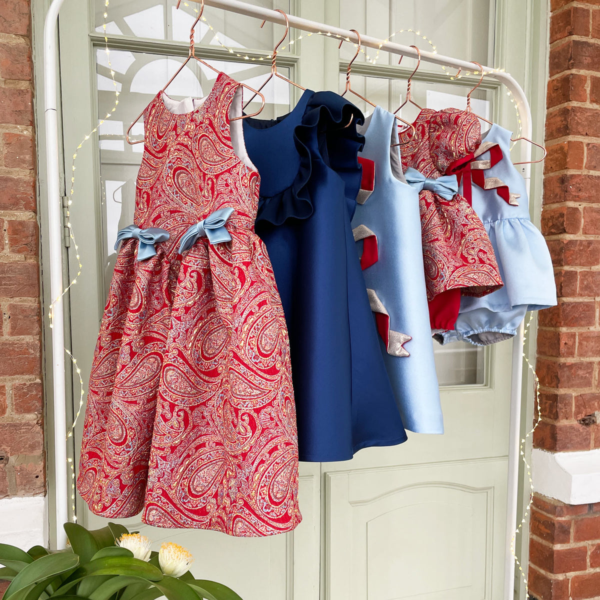 A set of four children's dresses and shorts hanging on a white clothing rack outdoors in front of a white door and brick wall. The set includes a red paisley dress with a blue bow, a solid navy blue ruffle dress, a light blue dress with red accents, and a light blue denim-style romper with a red bow.