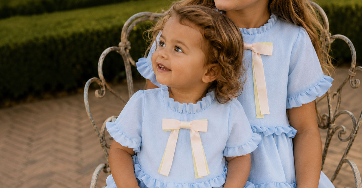 Two children in matching light blue dresses with bow details sitting on a decorative chair.